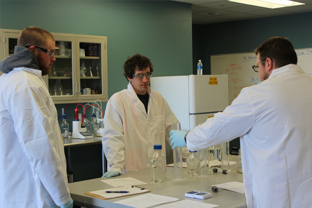 Three people in lab coats and gloves stand at a table.
